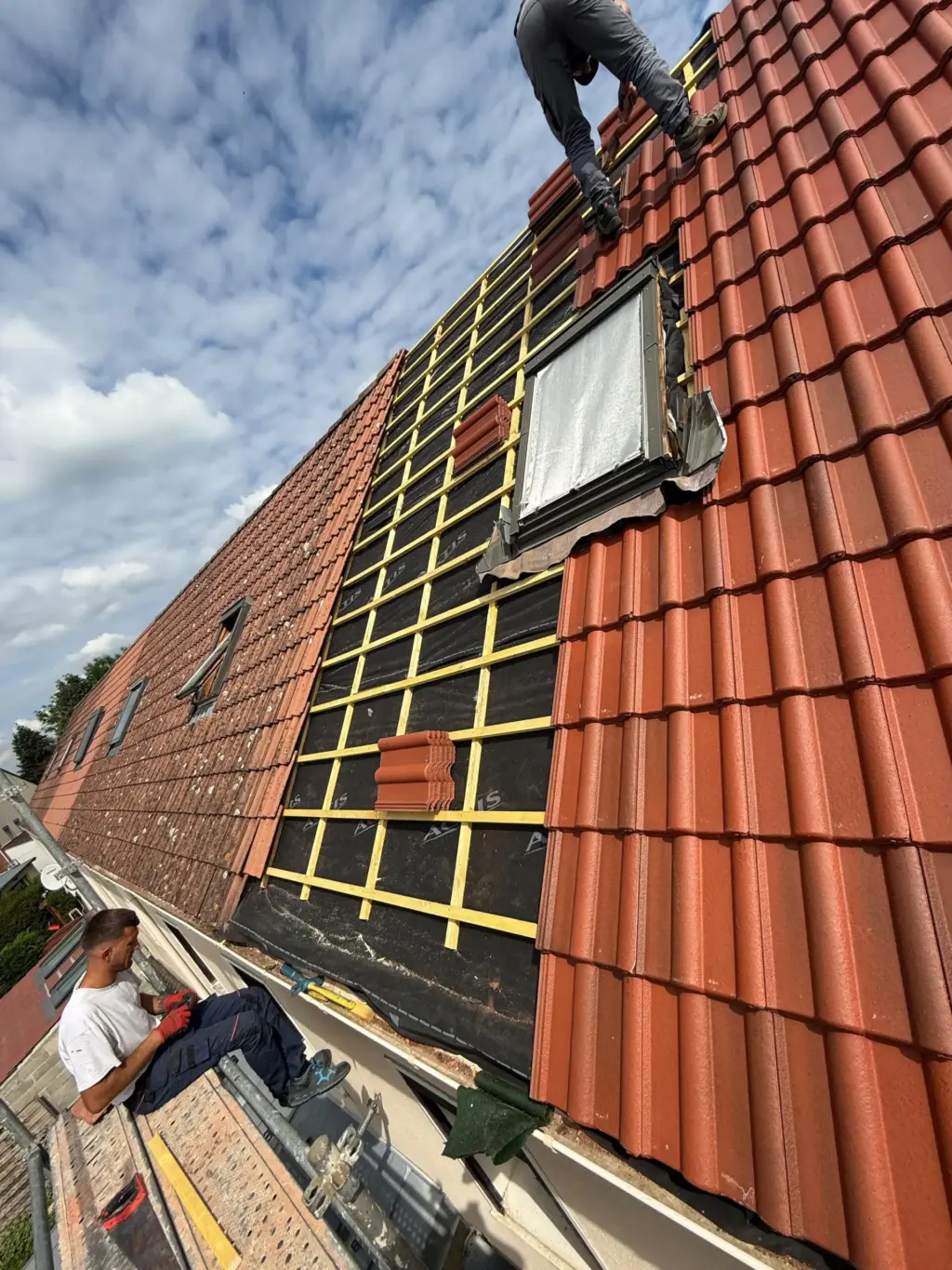 Workers installing red roof tiles on a sloped roof, with one person sitting on scaffolding and another standing on the roof.